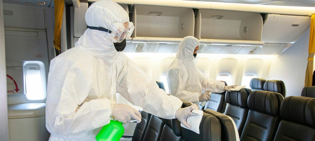 Two individuals in full protective gear sanitize the interior of an airplane, focusing on the seats and overhead compartments, offering valuable cleaning tips for maintaining a pristine environment.