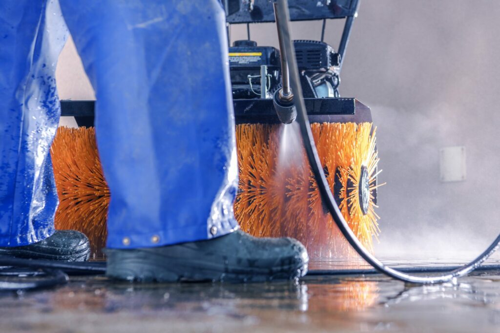 A person in blue waterproof gear, an expert in cleanup for industrial grime, operates a pressure washer to clean a surface with an industrial floor cleaning machine equipped with orange brushes.