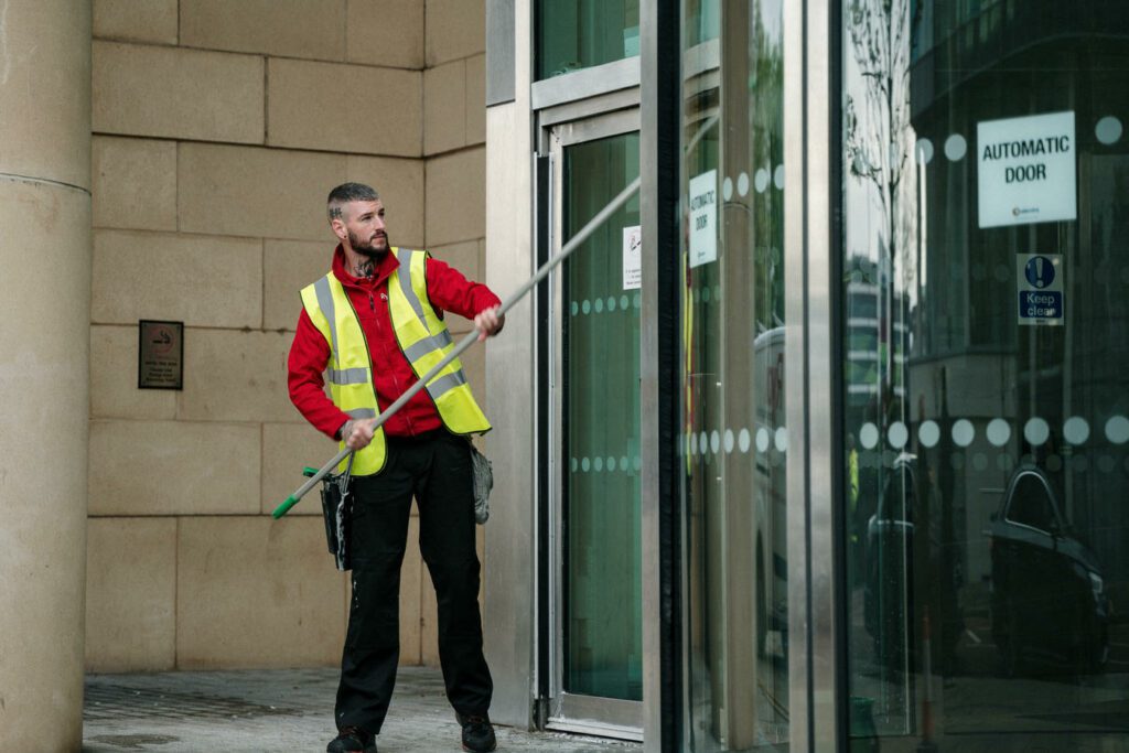 A person wearing a safety vest and red jacket is cleaning the exterior glass of a building's automatic door using a long-handled tool.