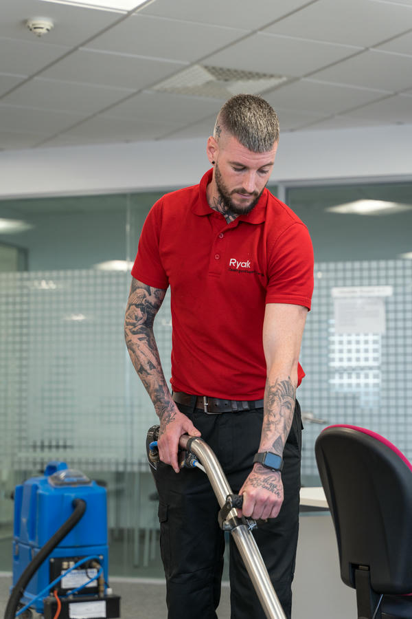 A man in a red shirt uses a vacuum cleaner in an office with gray partition walls and a blue cleaning machine in the background.