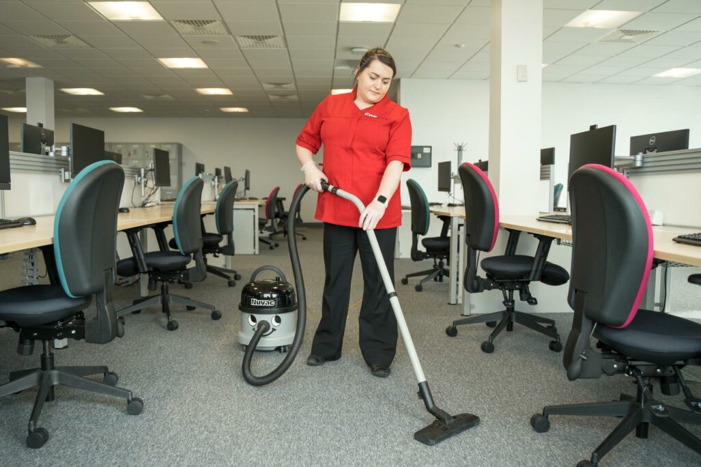A person wearing a red shirt is vacuuming the carpet in an office with multiple workstations and chairs.