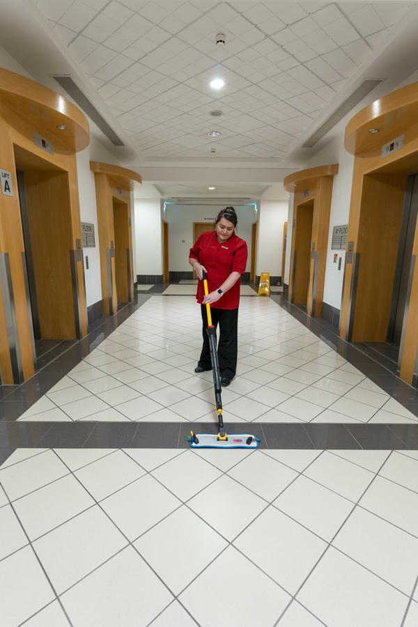 A person in a red shirt mops the tiled floor in a clean, well-lit hallway with four elevators.