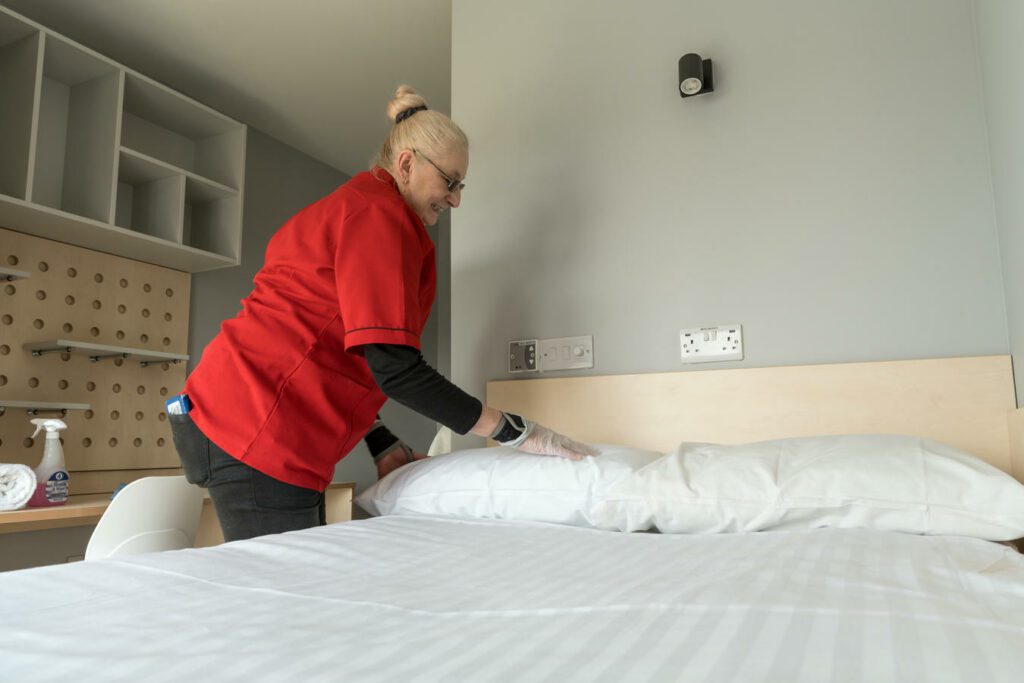 A person wearing a red shirt and gloves is making a bed in a clean, modern room with shelves and cleaning supplies visible.