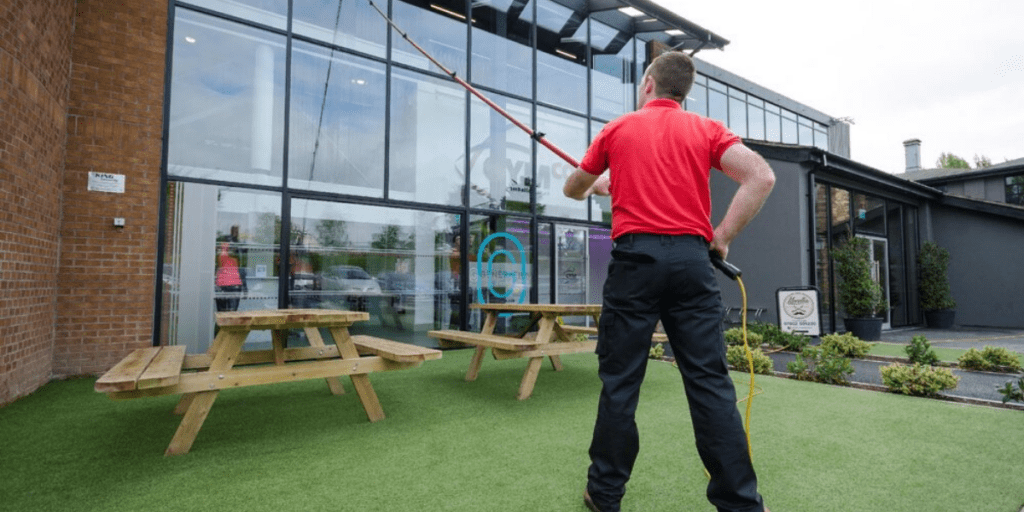 A man in a red shirt is performing commercial window cleaning on the exterior windows of a building in Belfast, using a long pole on a grassy area with picnic tables.