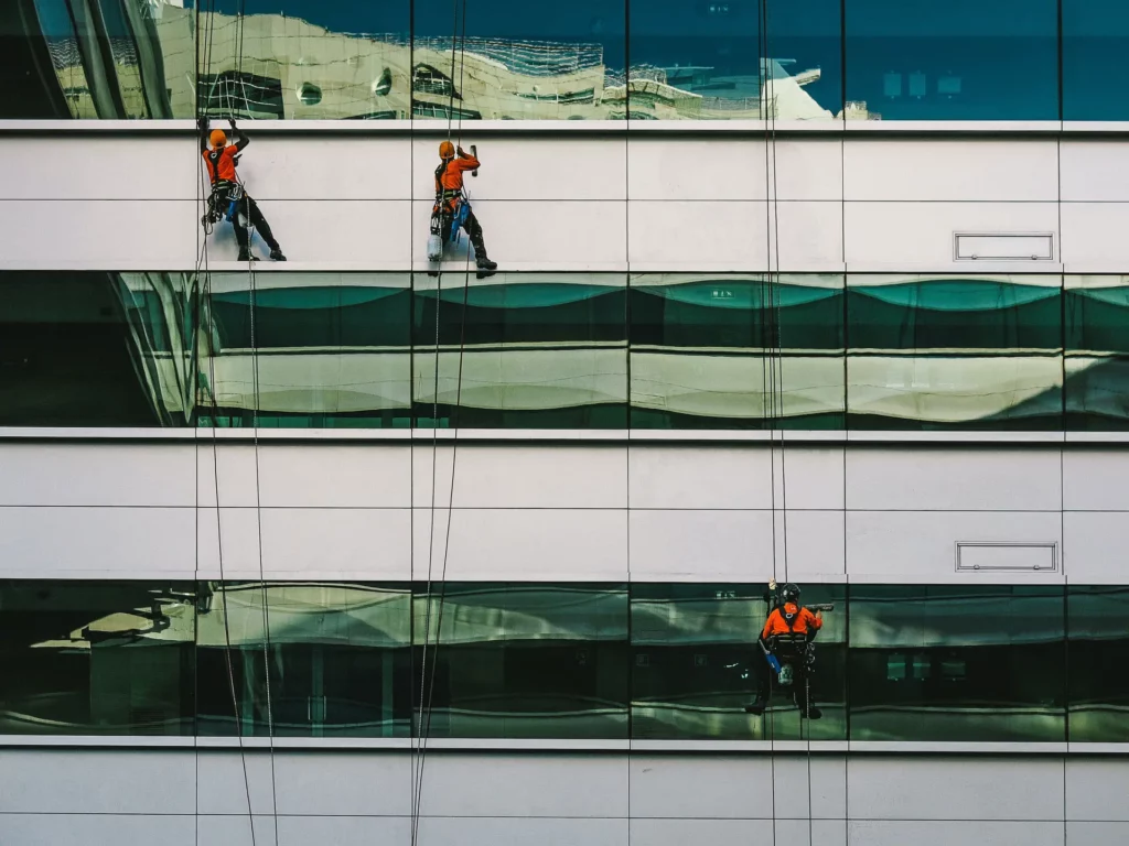 Three window washers with safety harnesses abseil down the side of a multi-story building, expertly performing window cleaning.