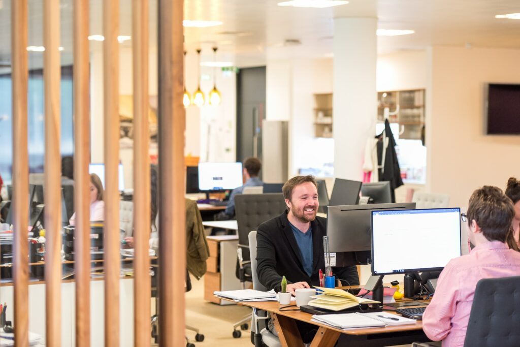People are working at desks with computers in a modern, open-plan office; a man is smiling and talking to a colleague. The high morale in the office contributes significantly to increased productivity.