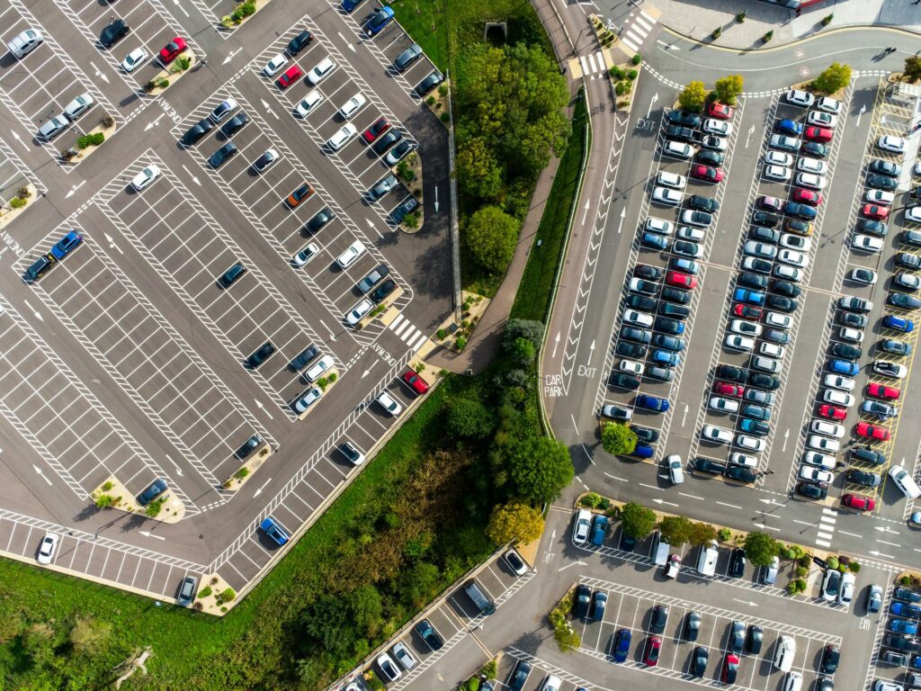 Aerial view of a large car park with numerous cars parked in designated spaces. The lot is divided into sections with marked lanes and pathways surrounded by greenery, providing a clear and organized layout ideal for regular cleaning services.
