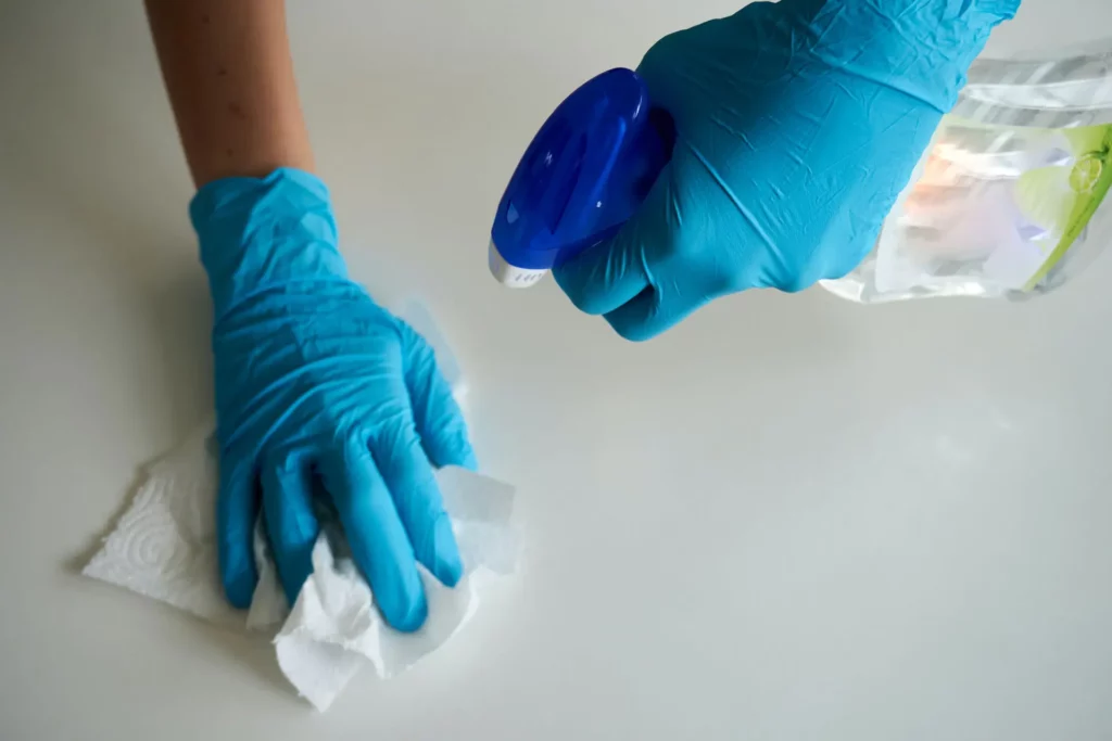 Two gloved hands are meticulously cleaning a white surface, one holding a spray bottle while the other wipes with a cloth, exemplifying expert facility cleaning techniques.