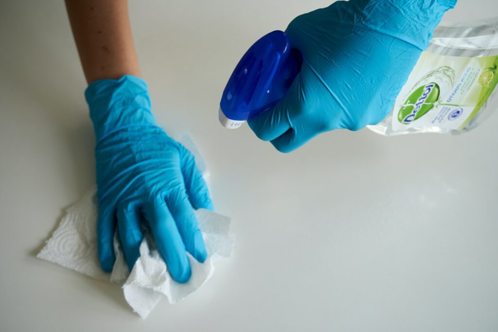 A person wearing blue gloves is performing periodic deep cleaning, diligently spraying a surface with a bottle and wiping it down with a paper towel.