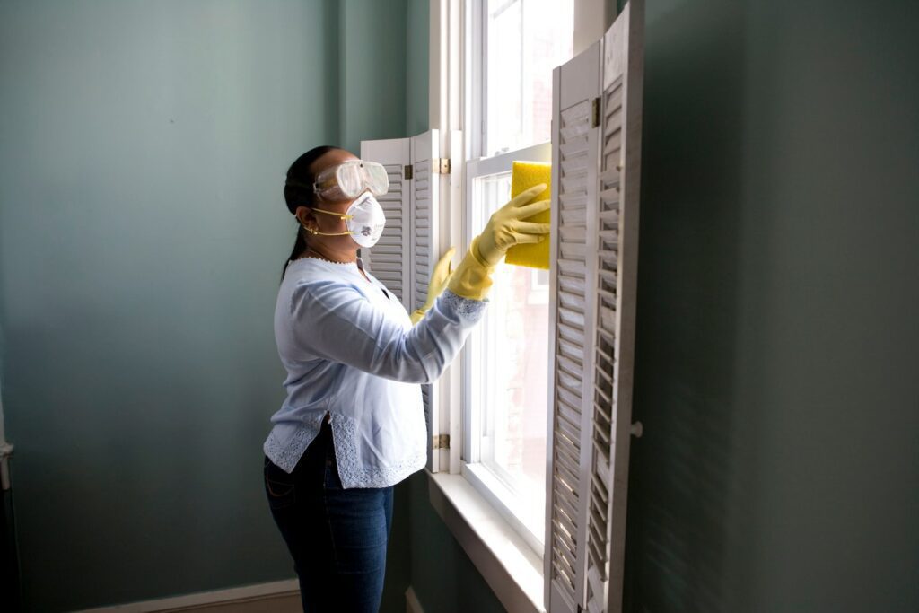 A person wearing protective goggles, a mask, and yellow gloves is cleaning a window with a sponge in a room with light blue walls. Imagine the benefits of hiring a professional cleaning company to get the job done right.