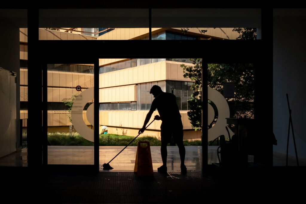 A silhouette of a person mopping the floor near a large glass entrance, with a caution sign in the middle, modern buildings visible outside, exemplifies top-notch janitorial services.