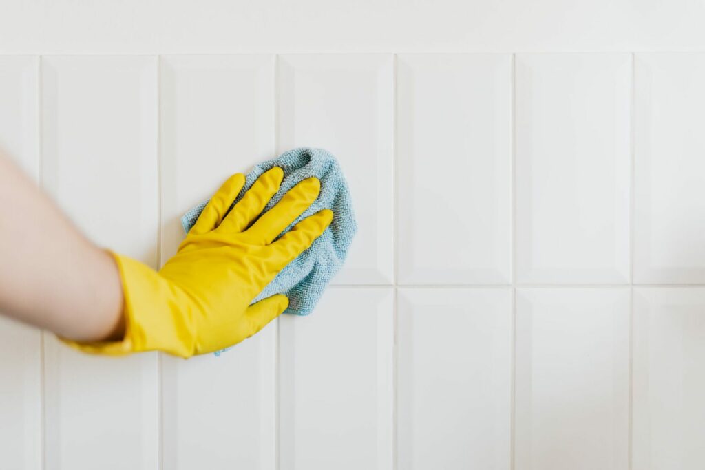 A hand wearing a yellow glove is cleaning a white tiled wall with a blue cloth, demonstrating the precision and care you can expect when you hire a cleaning service.