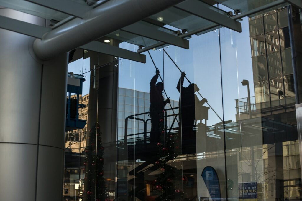 Two workers cleaning the exterior windows of a modern glass building using long poles, silhouetted against the glass under a clear sky.