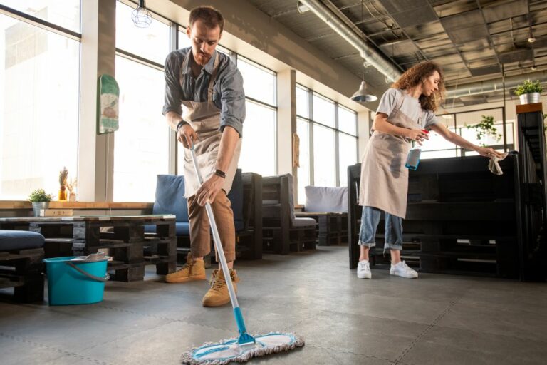 Two people in aprons are cleaning a commercial space. One is mopping the floor, and the other is wiping a surface.