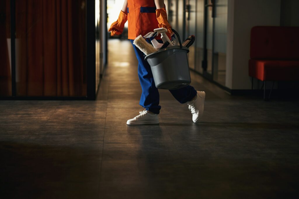 Person dressed in work attire, carrying a bucket filled with cleaning supplies, walking through a dimly lit hallway as part of the facility management team.