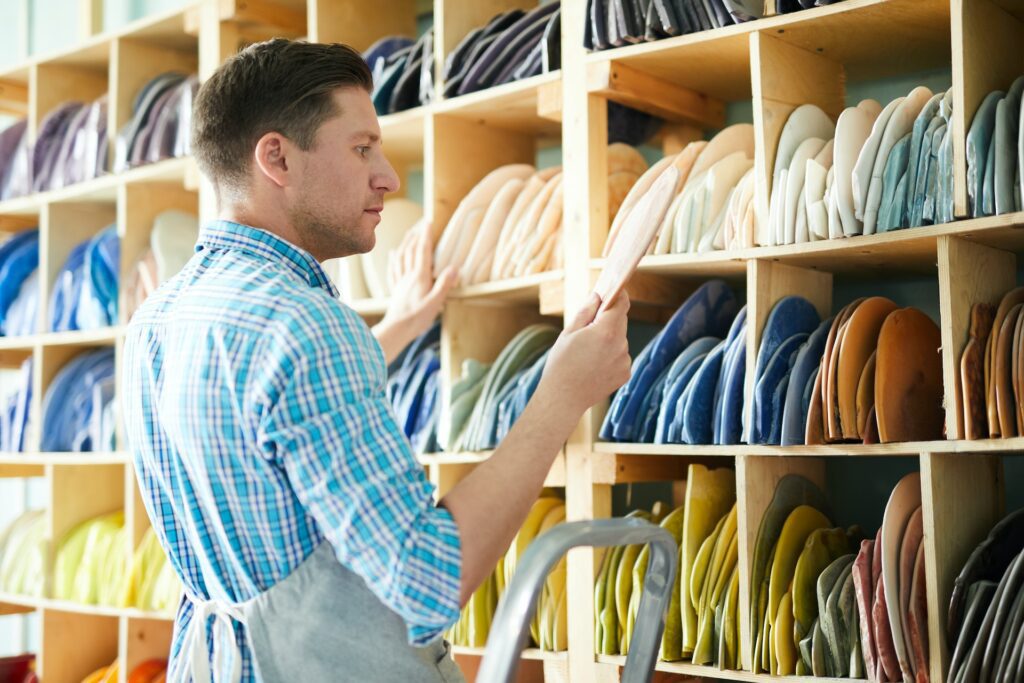 A person in a checked shirt and apron examines ceramic plates on a shelf filled with various colored plates.