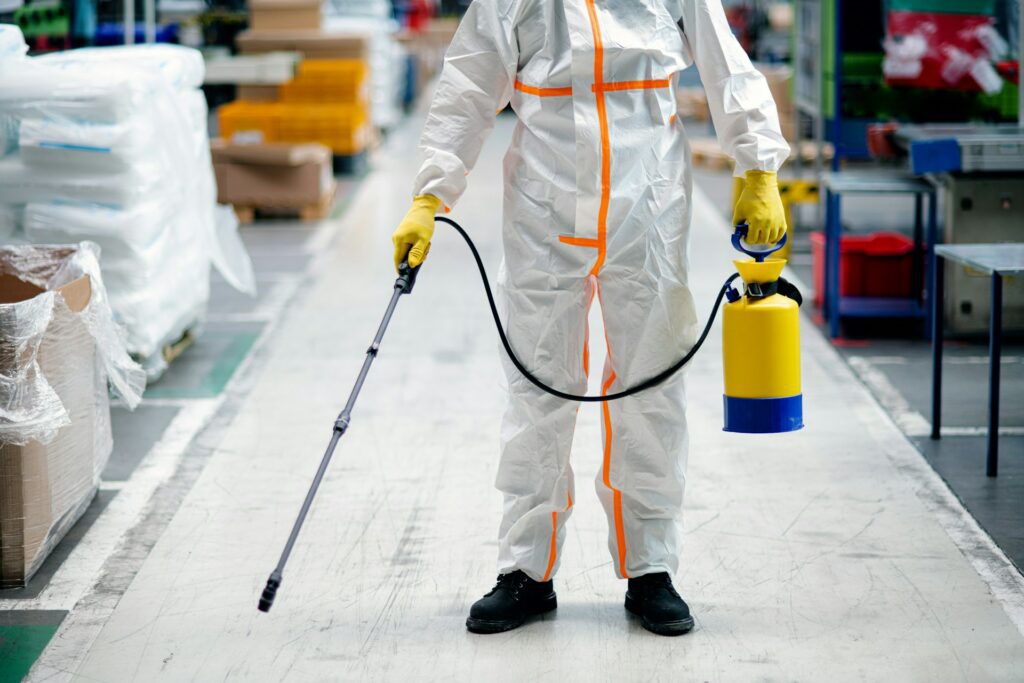 Person in protective suit and yellow gloves holding a sprayer in an industrial setting, ensuring a safe workspace.