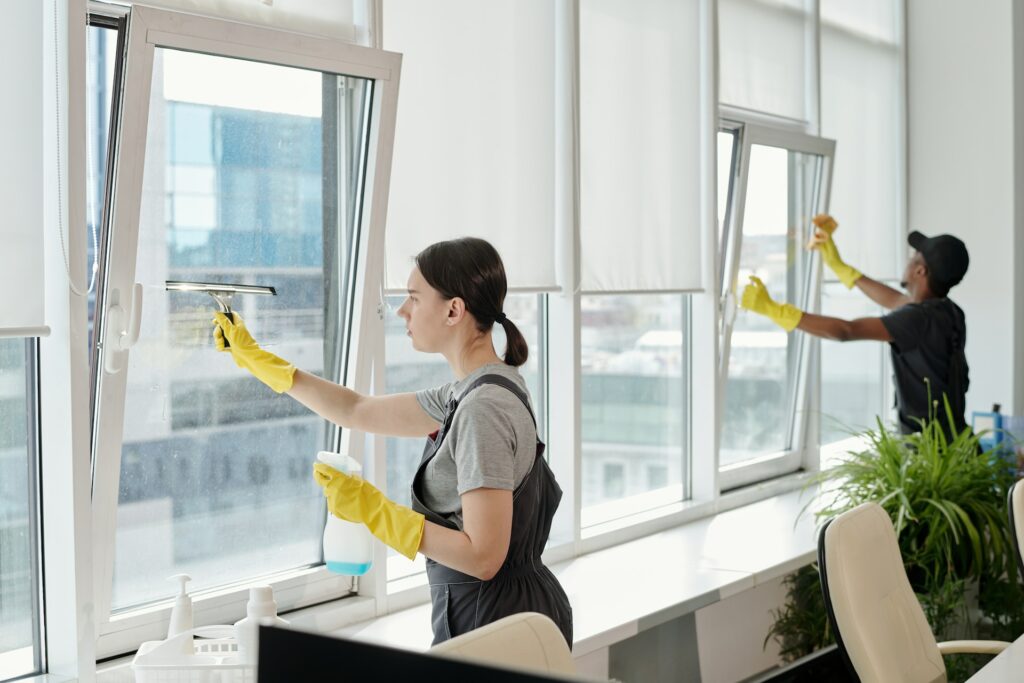 Two workers wearing gray uniforms and yellow gloves are cleaning office windows with cleaning supplies. A potted plant is on the windowsill. Office furniture is visible in the foreground.