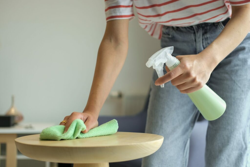 A person wearing jeans and a striped shirt cleans a round wooden table with a green cloth and spray bottle, showcasing the efficiency of commercial cleaning services.