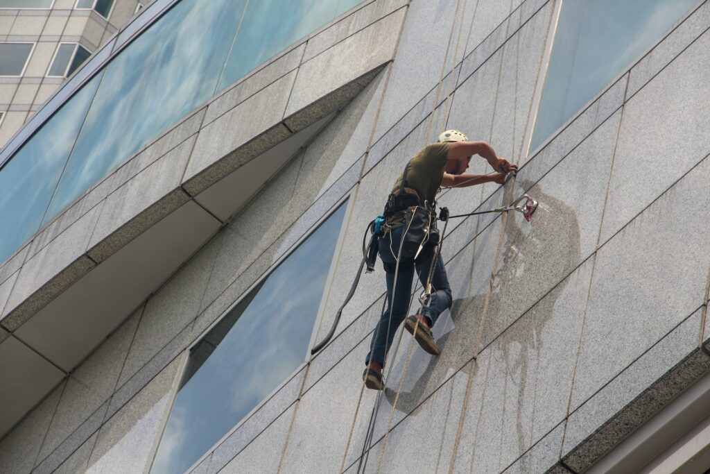 A worker in safety gear is suspended on the side of a glass building, using ropes and equipment to clean the windows, highlighting the importance of commercial window cleaning today.