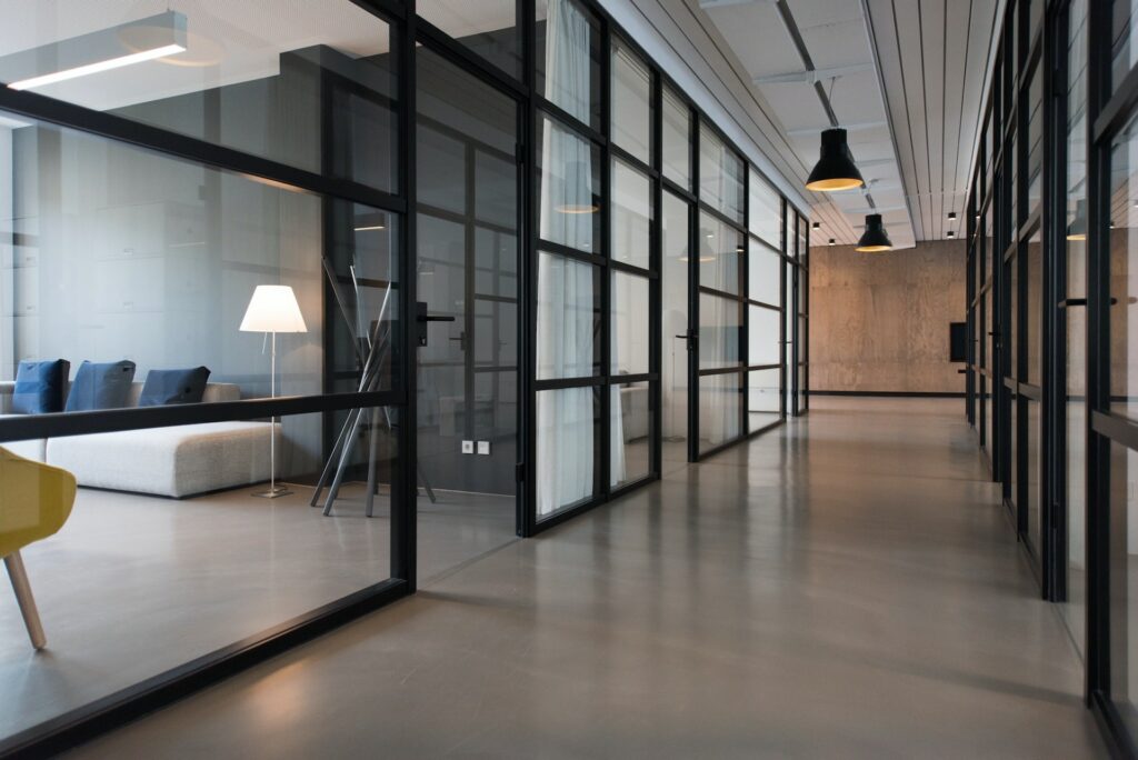 A modern office hallway with glass-walled rooms, a gray floor, and black-framed doors. A sofa, lamp, and chairs are visible inside one of the rooms. Black pendant lights hang from the ceiling.