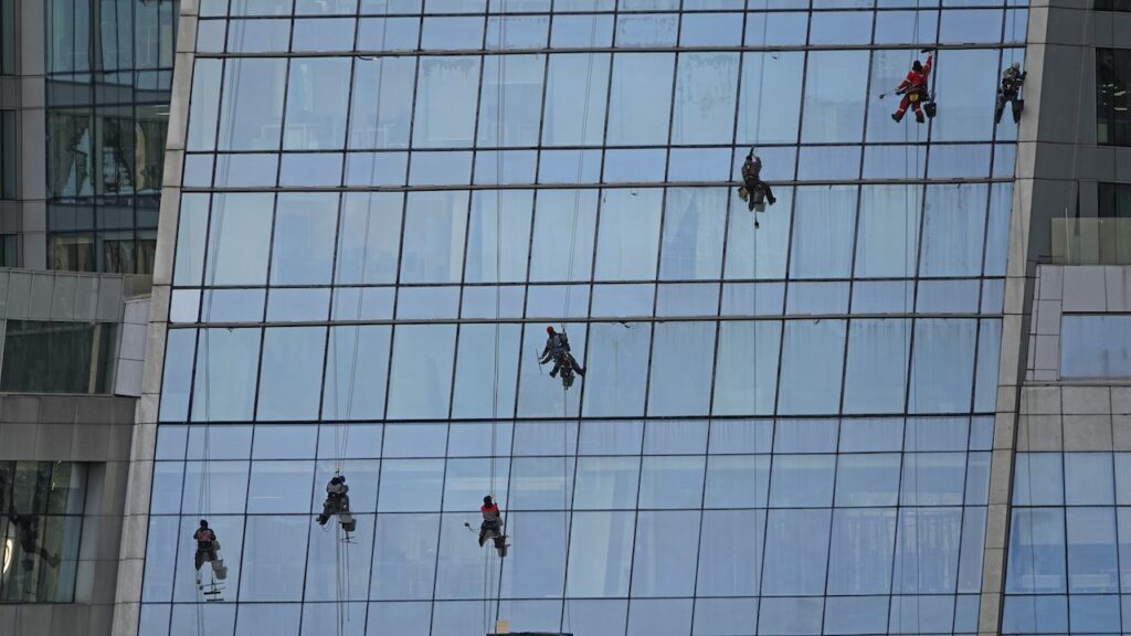 Window cleaners suspended from ropes are working on a large glass building, cleaning the windows from top to bottom.