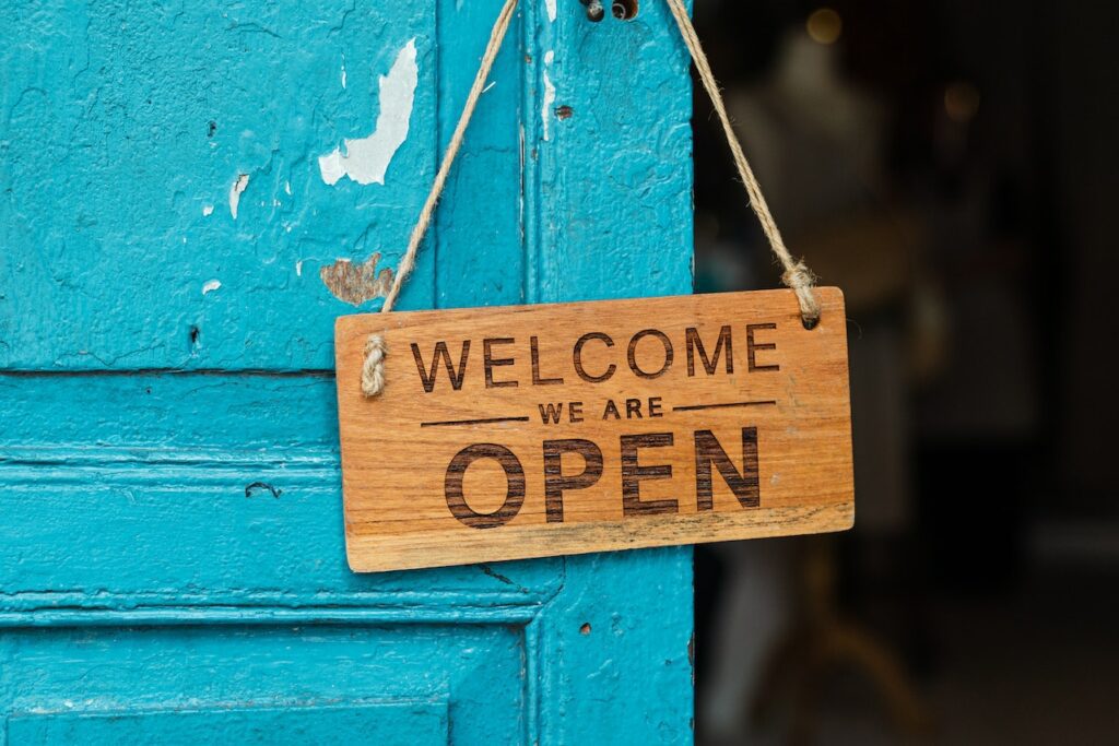 A wooden sign hanging on a blue door reads "Welcome, we are open.