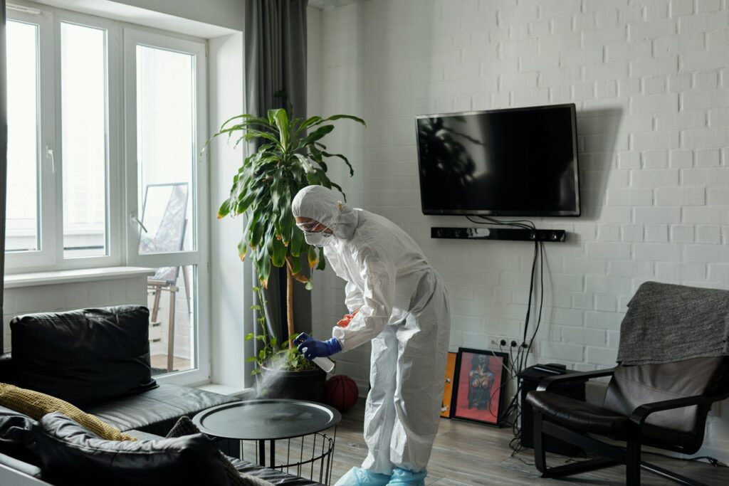 A person in full protective gear deep cleans a table in a modern living room with a TV on the wall and a large indoor plant, demonstrating why you need them for optimal hygiene.