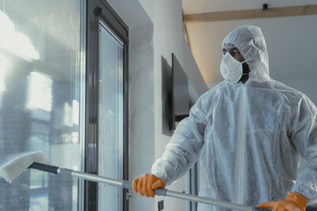 A person in a protective suit, gloves, and mask is providing professional commercial window cleaning services, meticulously mopping a glass door inside a building.