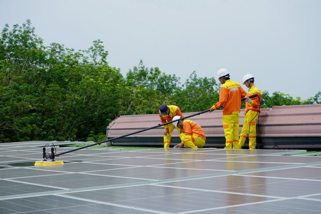 Four workers in safety gear installing or inspecting solar panels on a roof with greenery in the background.