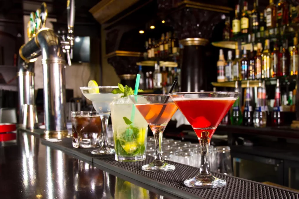 A bar counter at night clubs with an assortment of mixed drinks in various glasses, including cocktails and whisky, framed by a background of liquor bottles and taps.