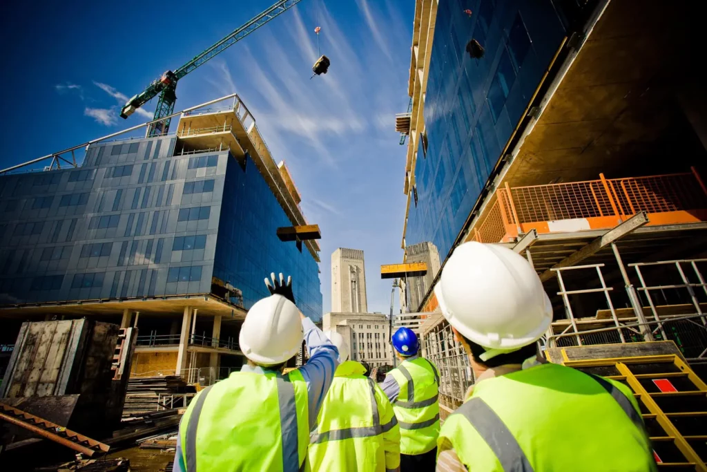 Construction workers in high-visibility vests and helmets observe a crane lifting materials between two partially constructed buildings under a clear blue sky, while a construction cleaning crew ensures the site remains tidy and safe.