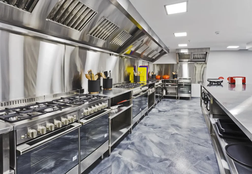Commercial kitchen with stainless steel appliances, including multiple stoves, ovens, and ventilation hoods. Kitchen tools are organized by the stove. The floor has a patterned finish and is easy to maintain for regular deep kitchen cleaning.