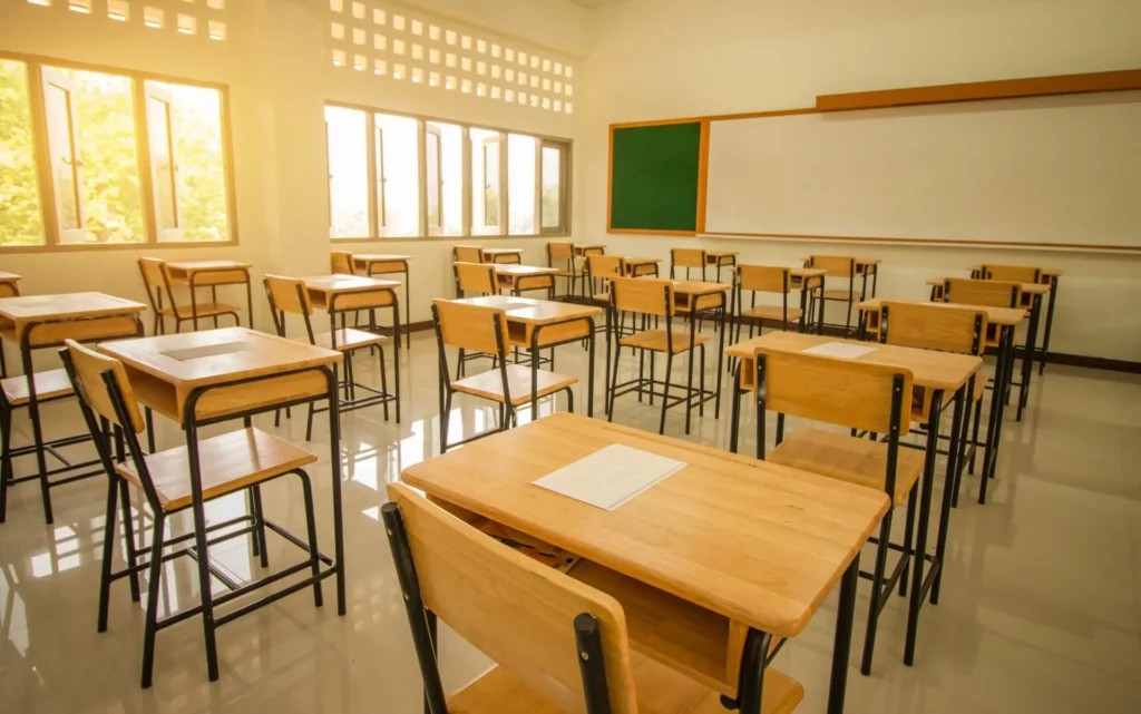 An empty classroom with rows of wooden desks and chairs, a chalkboard, and several large windows letting in sunlight, creating an inviting environment for education.