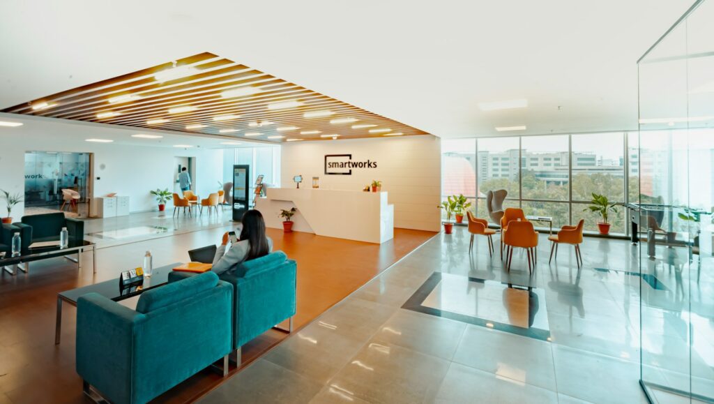 A modern office reception area with teal seating, a white front desk labeled "Smartworks," and a glass-walled meeting space with orange chairs in the background.