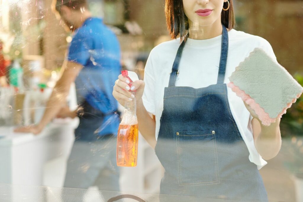 A woman in a white shirt and denim apron holds a spray bottle and a cleaning cloth, standing in front of a glass surface, ensuring retail cleanliness. Another person in a blue shirt is working in the background, contributing to customer satisfaction.