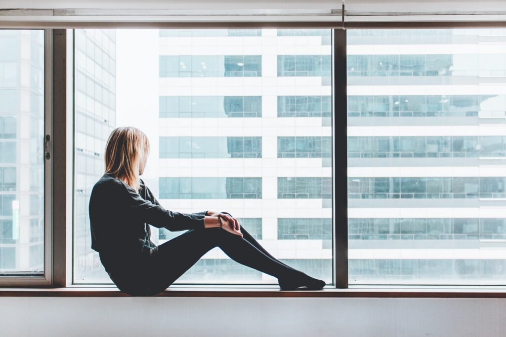 A person with long hair, dressed in dark clothing, sits on a windowsill looking out at the adjacent tall building through streaky windows.