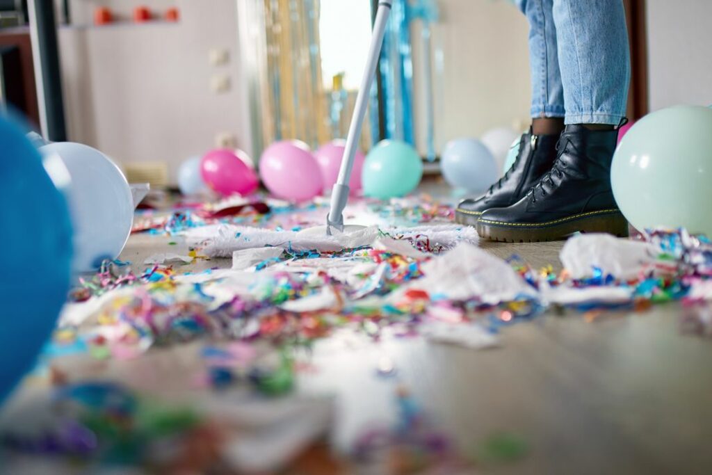 Person engaged in valuable afterparty cleanup, vacuuming confetti and balloons off the floor. Only legs in jeans and black boots are visible amidst the messy aftermath of what appears to have been a lively celebration.